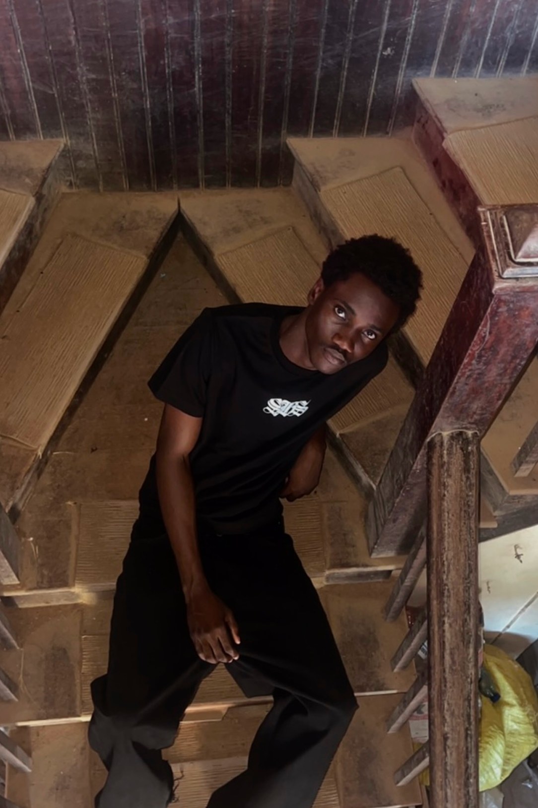 Kanann in a black tee, leaning against a wooden staircase