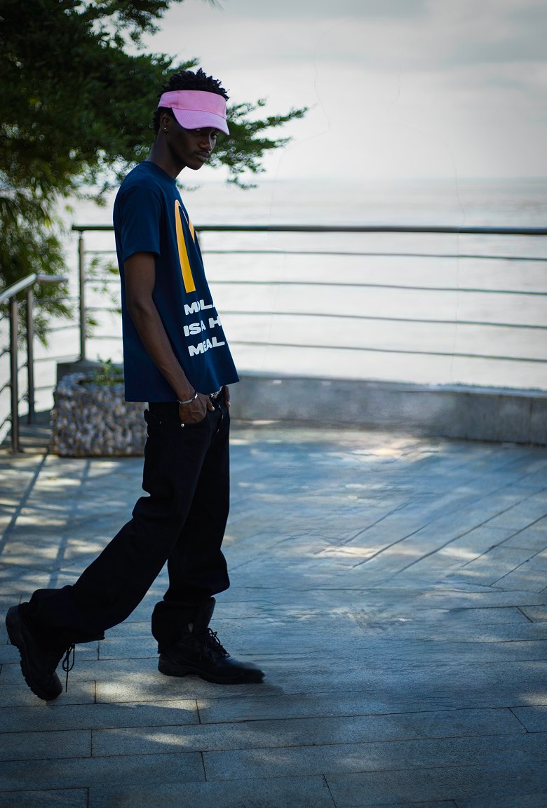 Kanann in a navy tee and black trousers, seen from the side on a seaside terrace