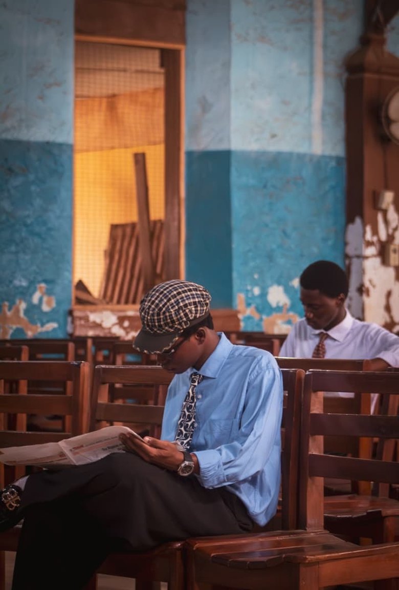 Kanann in a checked cap and blue shirt reading a newspaper on church pews