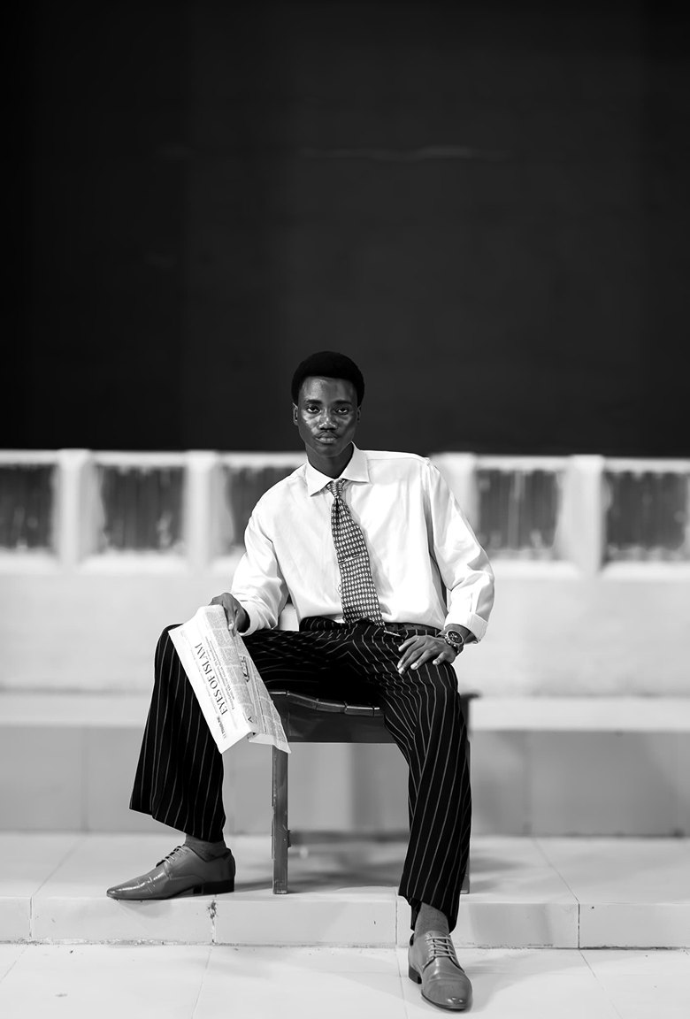 Black and white portrait of Kanann seated on a chair with a newspaper