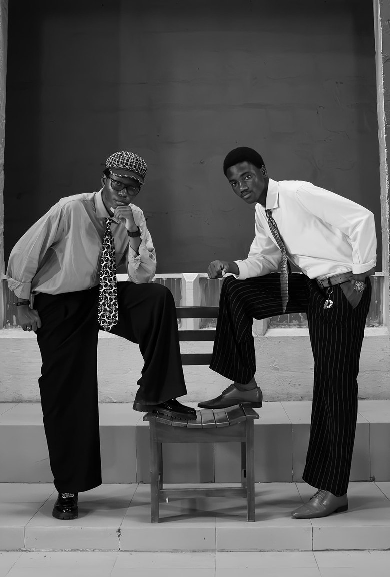 Black and white shot of two men in shirts, ties and pinstripe trousers around a chair