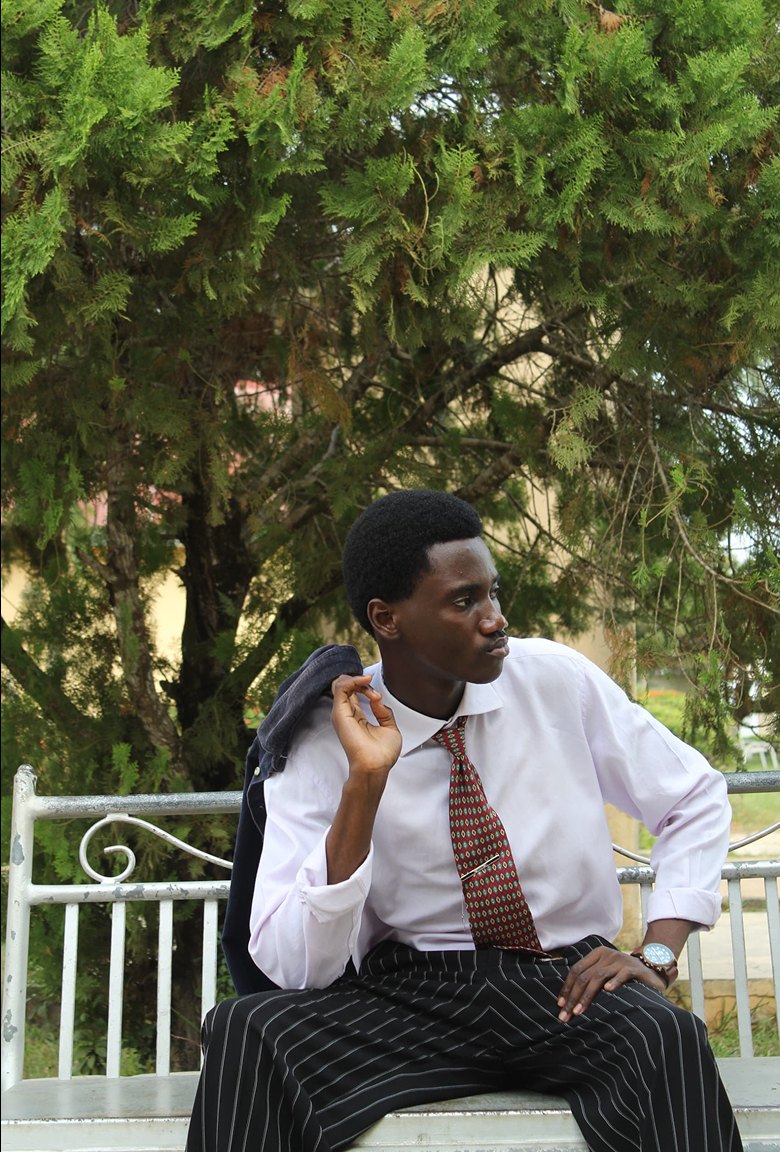 Kanann seated in a white shirt, red tie and pinstripe trousers