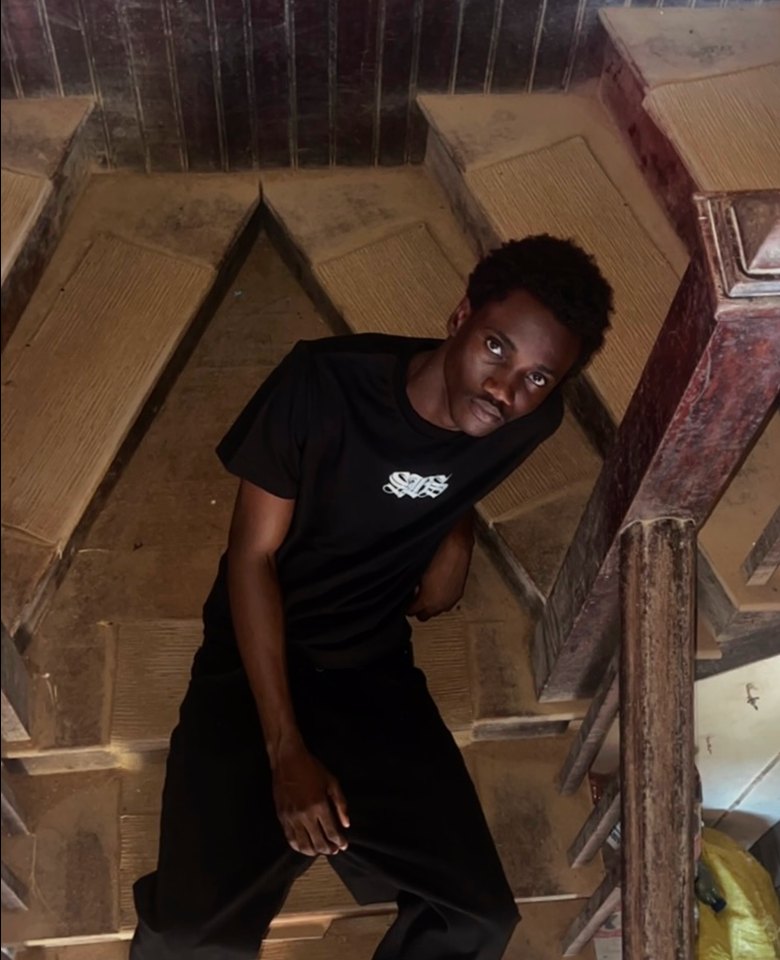 Kanann in a black tee leaning on a wooden staircase
